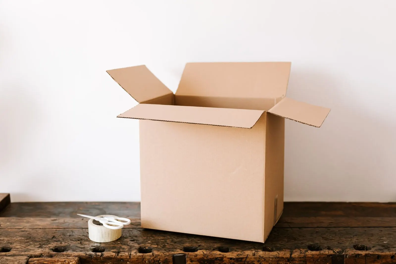 An open cardboard shipping box on a wooden desk, next to a roll of tape and scissors